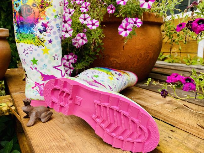 Gummistiefelpaar (ein Stiefel steht, der ander liegt so, dass man die Pinke Sohle sehen kann) auf einer Palette, im Hintergrund sind Blumen zu sehen. Die Gummistiefel sind bunt, mit vielen Farbflecken, Sternen und dem Hundemotiv Hazel, welches in Digitale