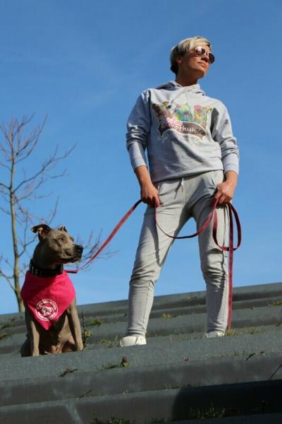 eine Frau steht auf Treppen und trägt einen grauen Jogginganzug mit dem Familienhunde-Logo von Rainbow Bridge Collection. Sie hält einen grauen Pitbull an der Leine, welcher ein pinkes Halstuchträgt. Der Himmerl ist blau und die Frau trägt eine Sonnenbril