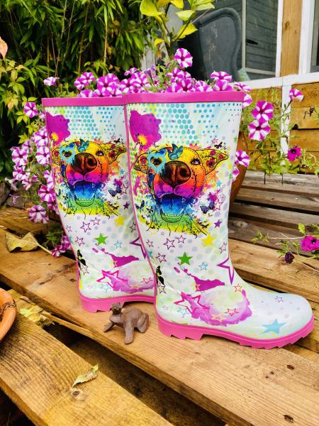 Gummistiefelpaar stehen neben einander auf einer Palette, im Hintergrund sind Blumen zu sehen. Die Gummistiefel sind bunt, mit vielen Farbflecken, Sternen und dem Hundemotiv Hazel, welches in Digitaler Popart gezeichnet wurde. Es ist komplett bunt und in